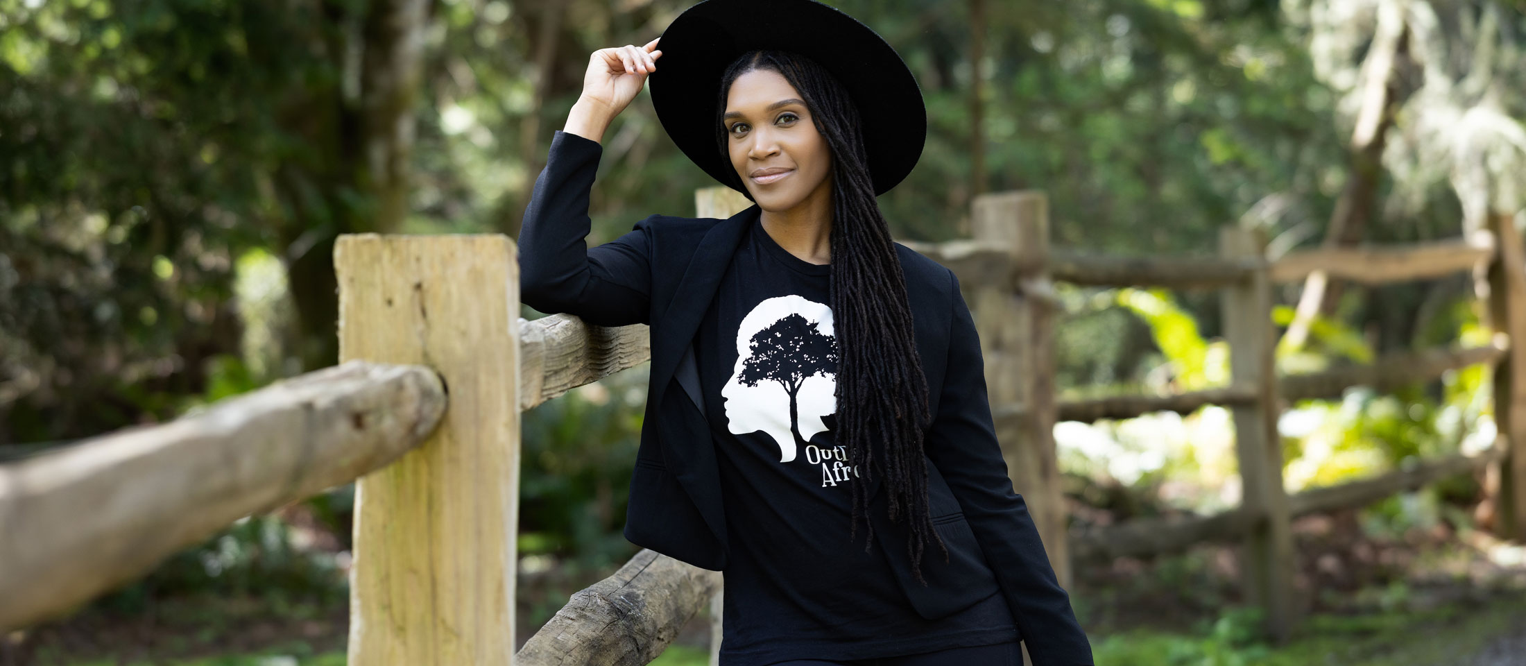 Woman wearing a black shirt and a black hat leaning against a fence with her hand on her hat, posing.